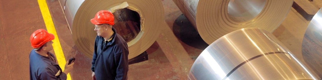 Two factory workers standing next to rolls of steel
