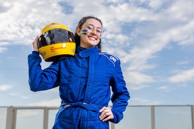 Mulher sorridente veste macacão azul de automobilismo e segura um capacete amarelo da Shell Eco-marathon Brasil sobre o ombro. Ela usa óculos e tem pinturas verdes e amarelas no rosto. Ao fundo, céu azul com nuvens.