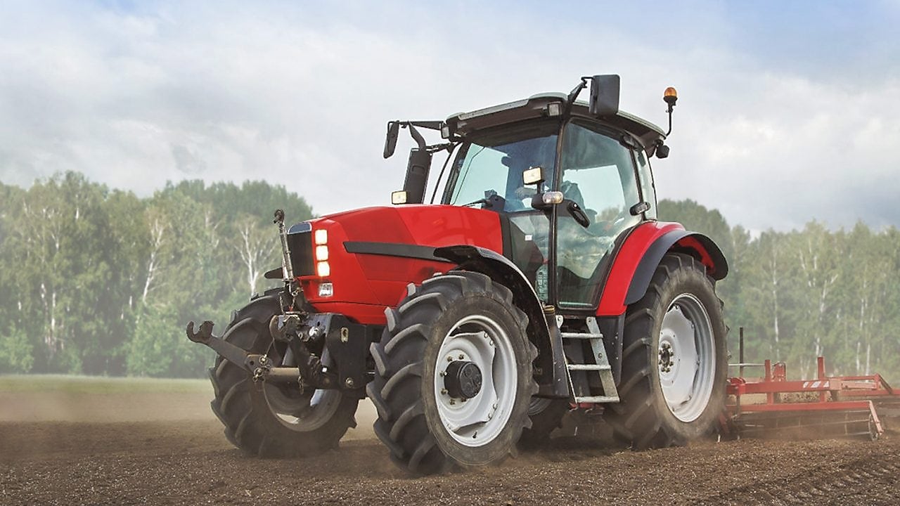 A red tractor ploughing a muddy field with trees in the background