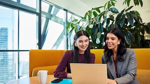 Two colleagues review content on a laptop in a bright office lounge