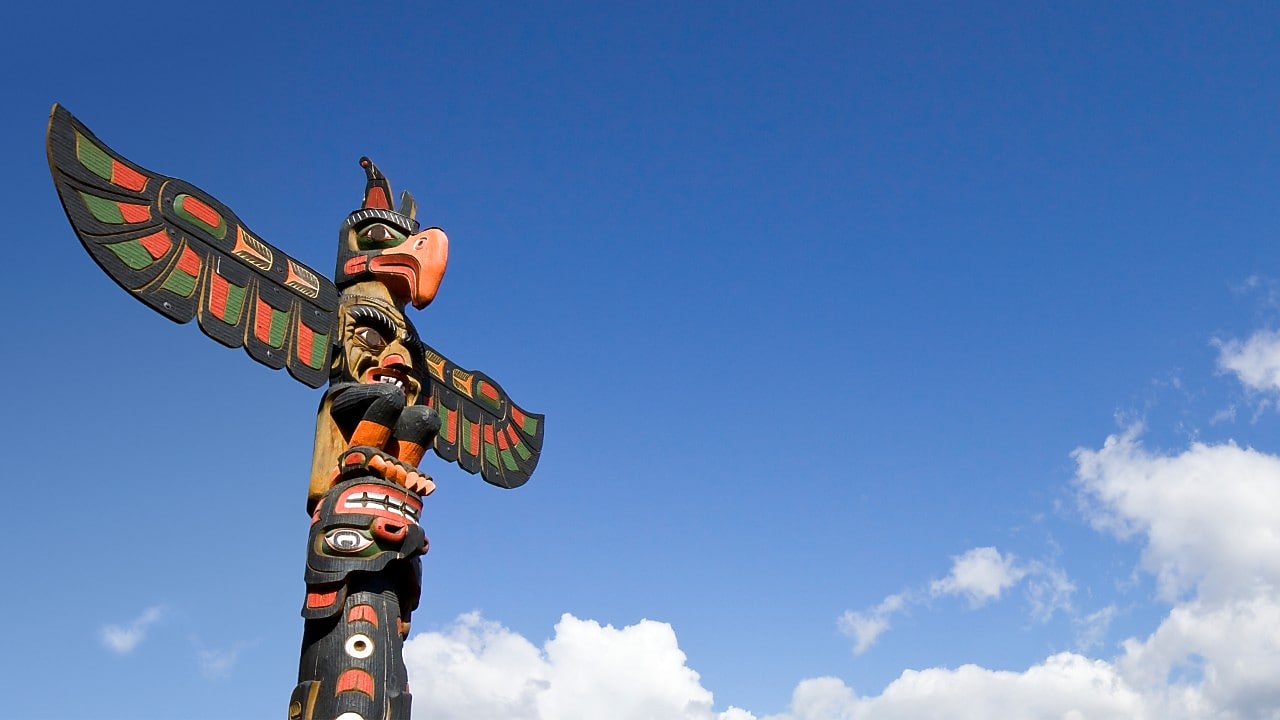 Vibrantly painted traditional totem pole featuring Indigenous carvings and a bird figure at the top, standing against a bright blue sky with scattered clouds.