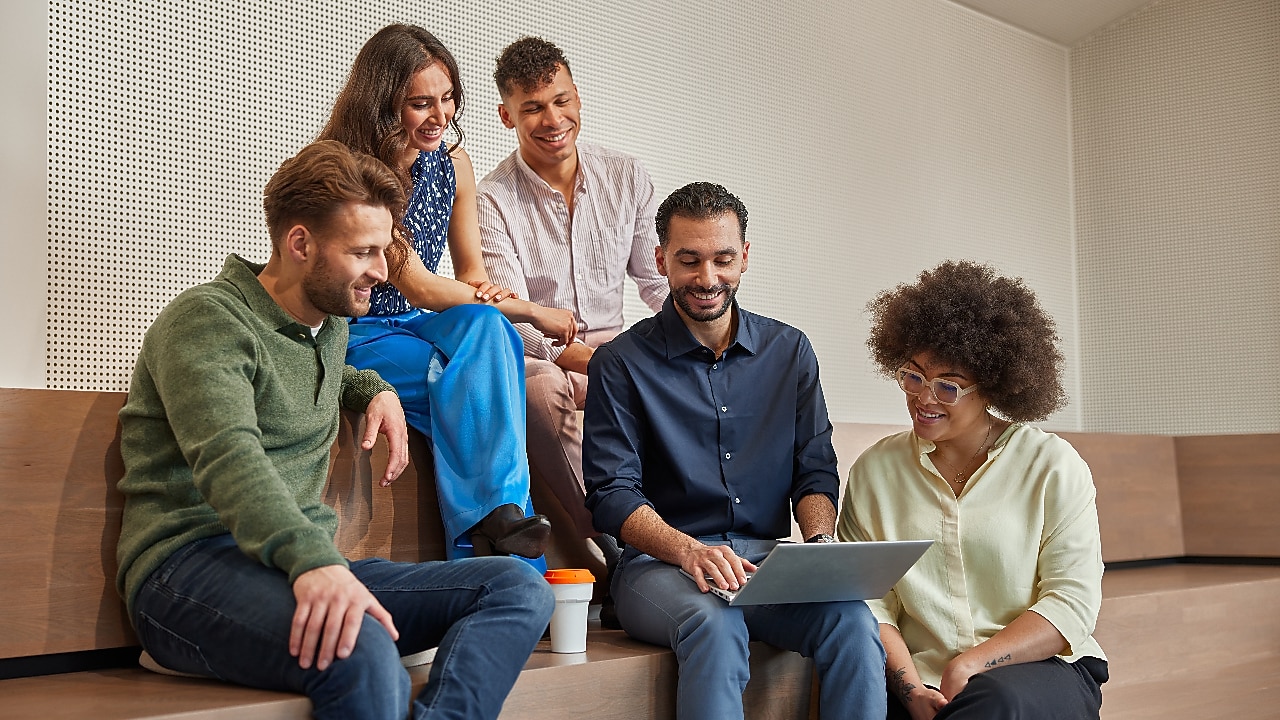 A group of five people are sitting on wooden benches in a modern, minimalistic room. They are gathered around a person using a laptop, with one person holding a coffee cup. The individuals appear to be engaged in a discussion or collaborative activity.