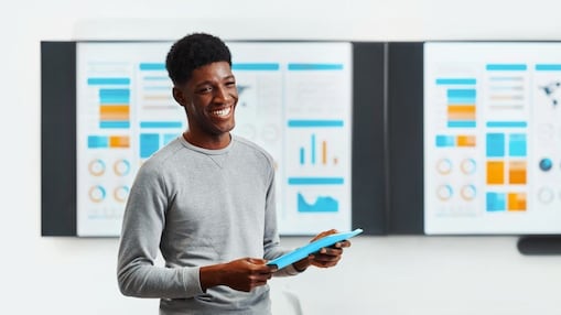 Professional presenting data on a tablet in a modern workspace, with large digital dashboards and charts displayed on screens behind them.