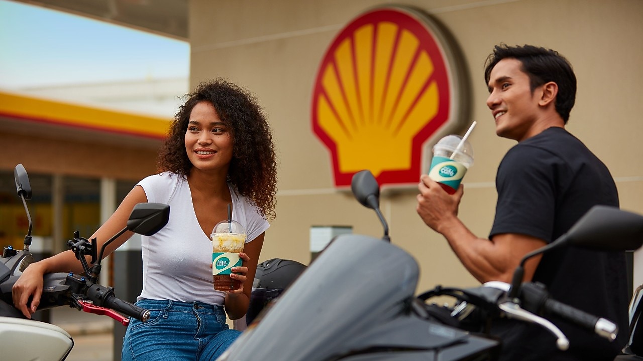 Two young customers enjoying Shell Café drinks outside a Shell service station with motorcycles.