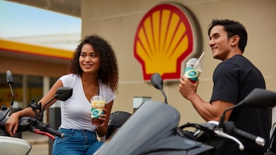 Two young customers enjoying Shell Café drinks outside a Shell service station with motorcycles.