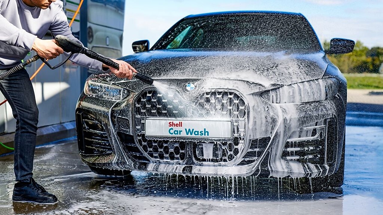 Person washing a black BMW car at a Shell Car Wash with soap and high-pressure water.