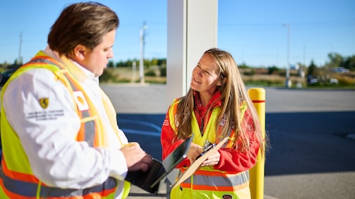 Two Shell service station employees in uniform smiling and greeting customers at the forecourt.