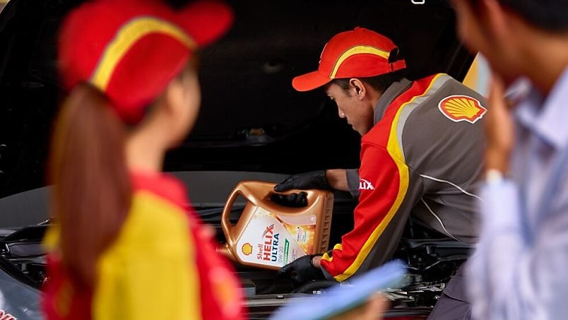 Shell mechanic pouring Shell Helix Ultra motor oil into a car engine while colleagues observe.