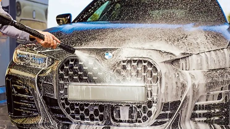 Close-up of a BMW car being washed with foam and a high-pressure water jet at a Shell car wash.
