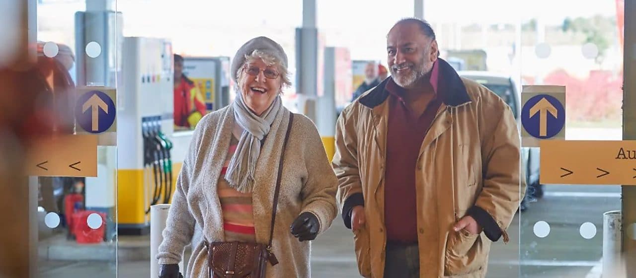 Two older customers smiling as they walk into a Shell service station store.