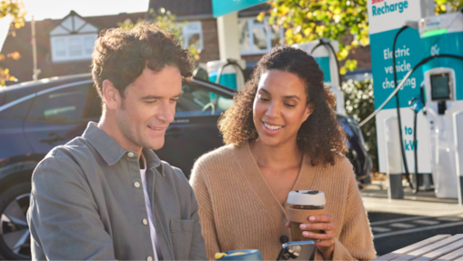 Two customers sitting at an outdoor table with coffee, using a smartphone while their electric vehicle charges at a Shell Recharge station.