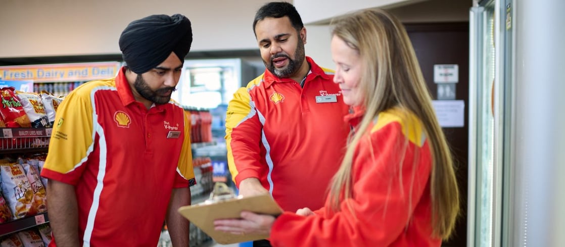 Shell service station team in red and yellow uniforms join hands in a show of teamwork and collaboration near fuel pumps.