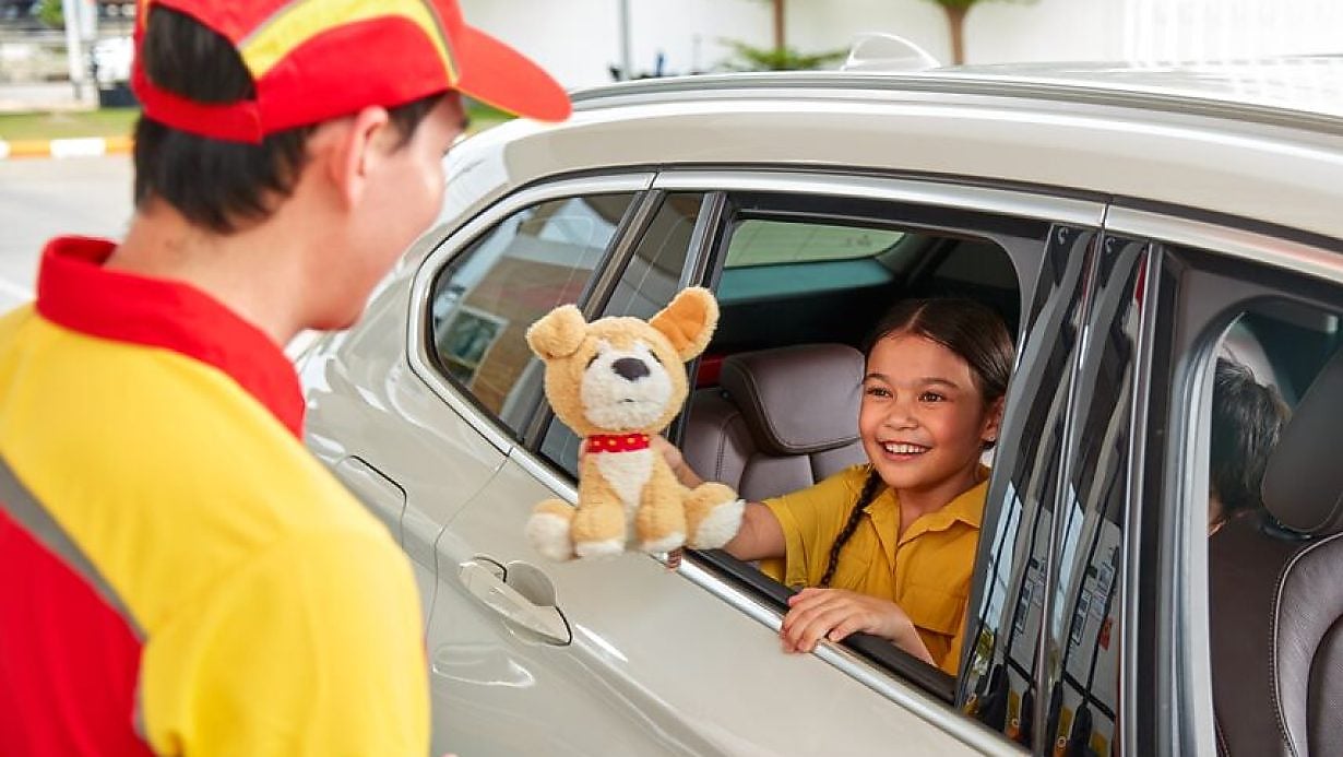 Child smiling and showing a stuffed toy to a Shell service station employee through a car window.