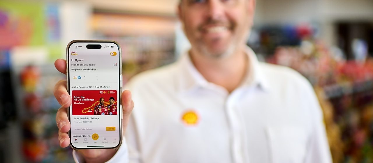 Person holding a smartphone showing the Shell Go+ app with a fuel rewards promotion, while drinking coffee outdoors.