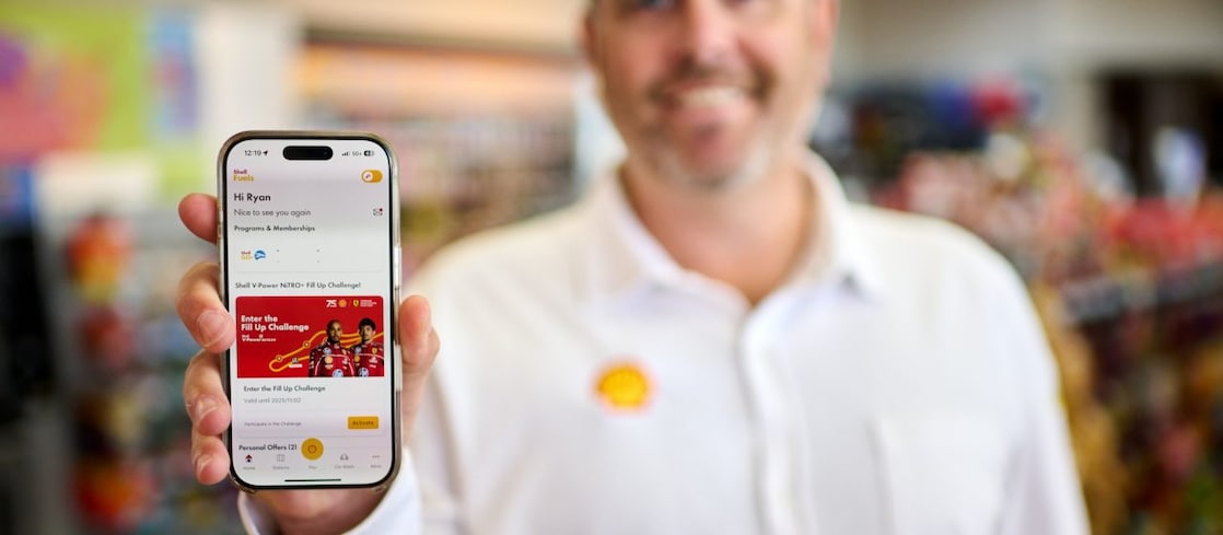 Person holding a smartphone showing the Shell Go+ app with a fuel rewards promotion, while drinking coffee outdoors.