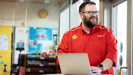 Shell employee in a red V-Power uniform smiling while working on a laptop inside a service station shop.