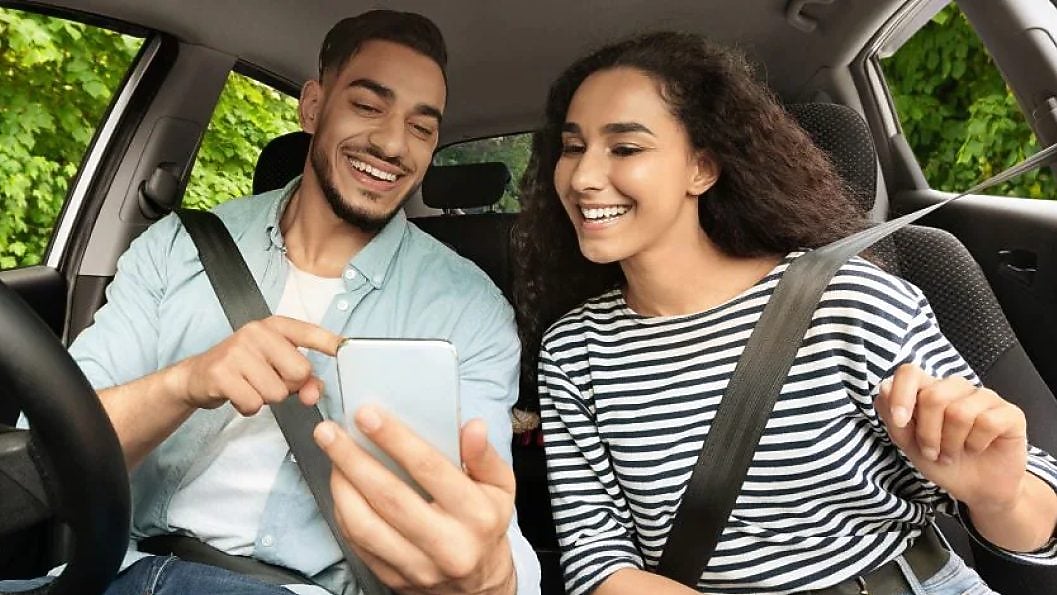 Two smiling young adults sitting in a car, looking at a smartphone together.