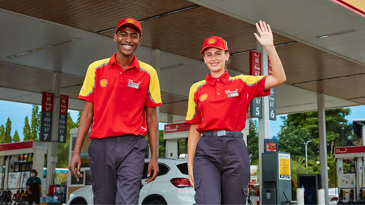 Two Shell service station employees in uniform smiling and greeting customers at the forecourt.