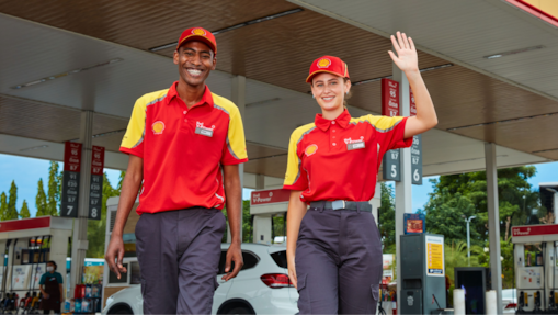 Two Shell service station employees in uniform smiling and greeting customers at the forecourt.