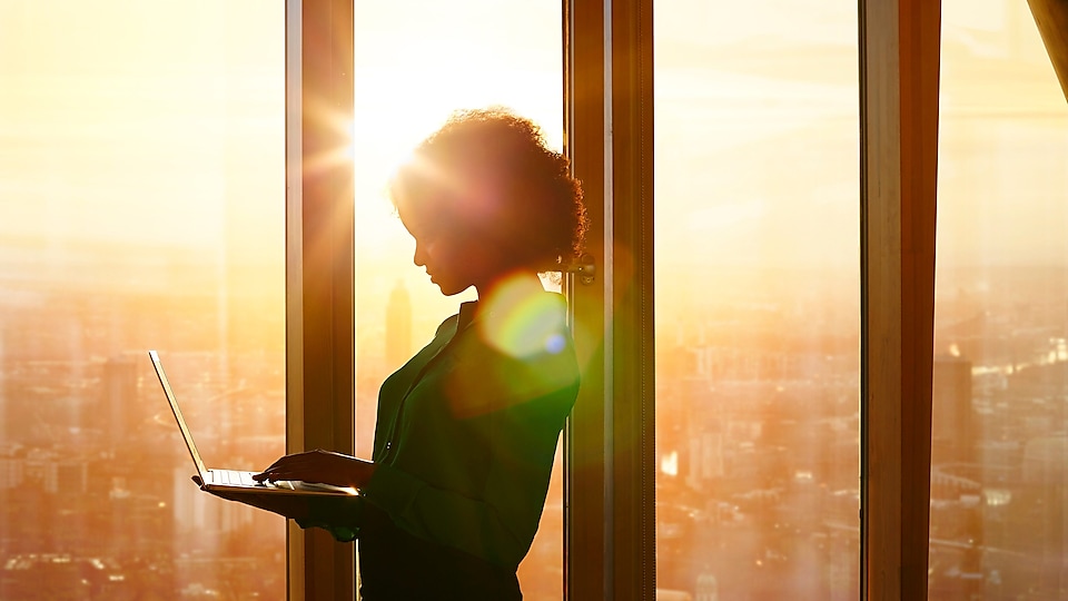 Une femme en position debout utilisant un portable dans un immeuble où l’on peut voir un coucher de soleil par les fenêtres