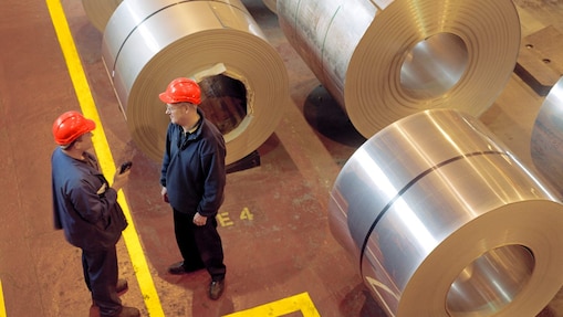 Two factory workers standing next to rolls of steel