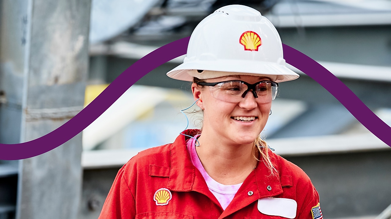 A person in a red Shell uniform and white hard hat with the Shell logo stands in an industrial setting.
