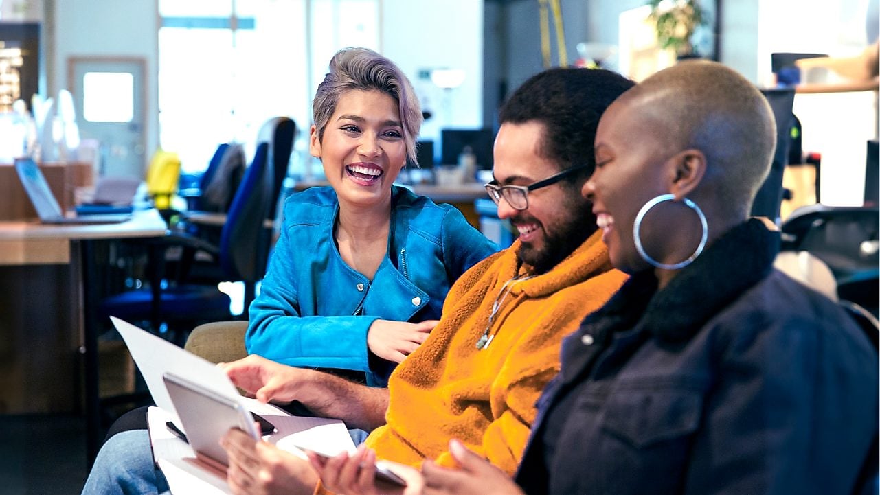 Three individuals are seated in an office setting, engaged in a discussion. The person on the left is wearing a blue jacket, the person in the middle is wearing an orange sweater, and the person on the right is wearing a dark jacket. They are holding papers and appear to be collaborating or reviewing documents. The background shows office furniture, including desks and chairs, with some computer monitors visible.