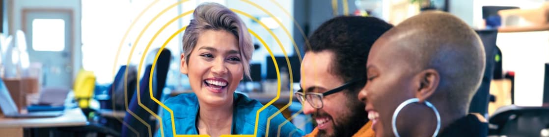 Three individuals are seated in an office setting, engaged in a discussion. The person on the left is wearing a blue jacket, the person in the middle is wearing an orange sweater, and the person on the right is wearing a dark jacket. They are holding papers and appear to be collaborating or reviewing documents. The background shows office furniture, including desks and chairs, with some computer monitors visible.