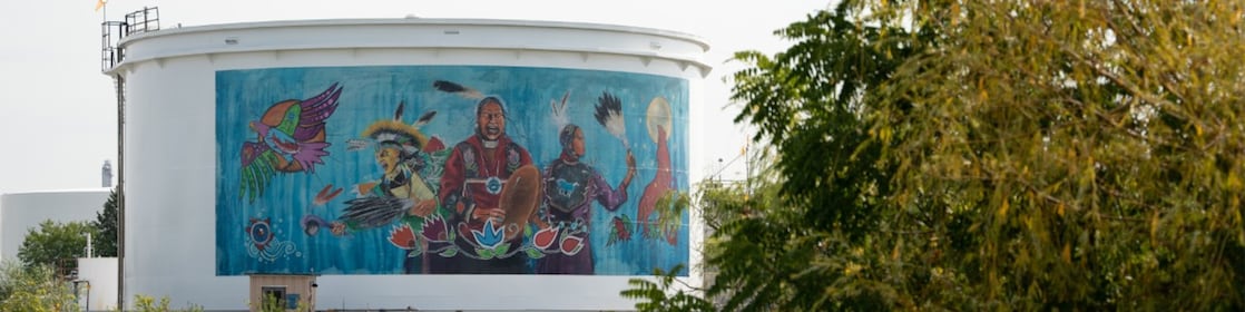 Peinture murale sur un grand réservoir d’eau représentant trois Autochtones en tenue traditionnelle, avec une figure centrale tenant un tambour, entourée d’oiseaux, de plumes et de plantes indigènes, symbolisant la culture et le patrimoine autochtones.
