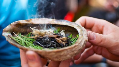 Close-up of hands performing a smudging ritual with a smoking shell containing pine needles, herbs, and ash, used in Indigenous spiritual practices.