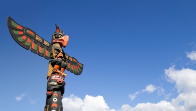 Vibrantly painted traditional totem pole featuring Indigenous carvings and a bird figure at the top, standing against a bright blue sky with scattered clouds.