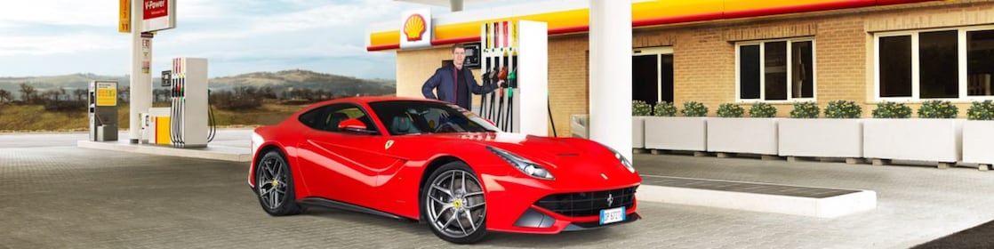 Ferrari rouge stationnée devant une station d'essence Shell avec un homme adossé à une pompe à essence